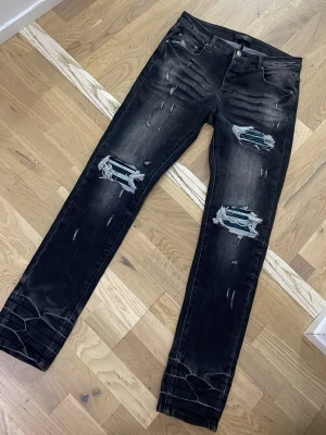 Svarta ripped jeans från Amiri - Säljer ett par svarta jeans från Amiri med slitna detaljer och stora hål på båda benen. Jeansen har fem fickor, knappgylf och en smal passform. Perfekta för dig som gillar en edgy och trendig stil.