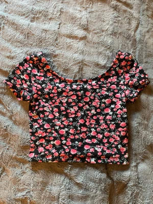 Söt sommar topp - Superfin croppad topp från Divided med svart botten och blommigt mönster i rosa och vitt. Toppen har korta ärmar och en rundad halsringning. Perfekt att styla med jeans eller kjol för en trendig look.