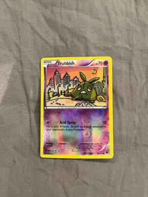 Pokémonkort Trubbish holo - Säljer ett Pokémonkort med Trubbish-motiv. Kortet har en färgglad holo-effekt och gul ram. Motivet visar Trubbish framför en stadssilhuett i solnedgång. Text och detaljer på engelska. Perfekt för samlare eller Pokémonfans.