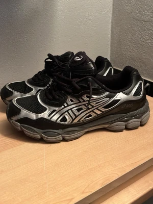 Svarta och silver Asics sneakers - Snygga sneakers från Asics i svart och silver med klassisk logga på sidan. Skorna har meshpaneler för bra ventilation och detaljer i syntetmaterial. Tjock sula med GEL-dämpning och snörning framtill för skön passform.