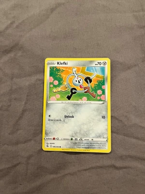 Pokémonkort Klefki - Säljer ett Pokémonkort med Klefki-motivet. Kortet har gul ram och färgglad illustration med Klefki och nycklar mot en grön och orange bakgrund. Text och symboler i svart och grått. Perfekt för samlare eller Pokémonfans.