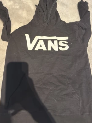 Svart hoodie från Vans med logga - Svart hoodie från Vans med stor vit logga på bröstet. Klassisk känguruficka framtill och justerbar huva med snören. Tillverkad i mjukt bomullsmaterial, perfekt för en avslappnad streetwear-look.