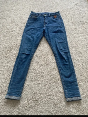 Blå skinny jeans  - Säljer ett par blå skinny jeans  med klassisk femficksdesign och uppvikta benslut. Jeansen har en snygg tvättad look och ett brunt läderpatch Perfekta för en trendig och avslappnad stil.