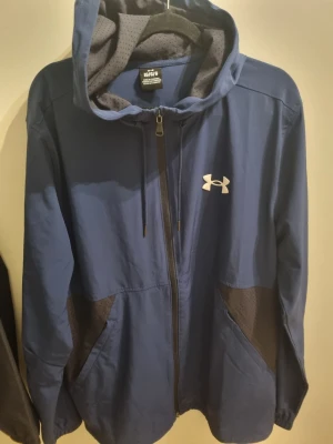 Blå vindjacka från Under Armour - Bllå vindjacka från Under Armour med dragkedja, huva och svarta mesh-detaljer på fickorna. Jackan har vitt logotryck på bröstet och ventilerande meshfoder i huvan.  Fast pris 450 kr
