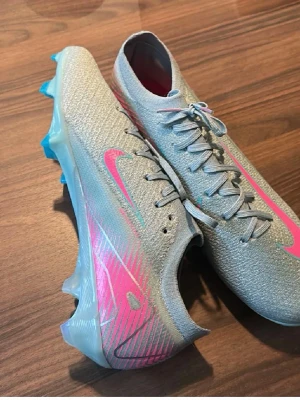 Nike Mercurial grå/rosa fotbollsskor - Snygga Nike Mercurial fotbollsskor i grått med rosa detaljer och blå dobbar. Skorna har en modern, strömlinjeformad design och är tillverkade i ett lätt syntetmaterial för bästa känsla och grepp på planen. Perfekta för dig som vill sticka ut på matchen.