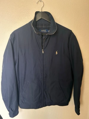 Blå vindjacka från Polo Ralph Lauren - Klassisk marinblå vindjacka från Polo Ralph Lauren med dragkedja framtill och broderad logga på bröstet. Jackan har ståkrage med kontrastfärgad insida och två snedställda fickor. Perfekt för en stilren och avslappnad look.