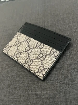 Gucci korthållare  - Snygg korthållare från Gucci i beige canvas med klassiskt GG-mönster och svarta läderdetaljer. Plats för flera kort på båda sidor. Perfekt accessoar för dig som gillar stilrena och lyxiga detaljer.