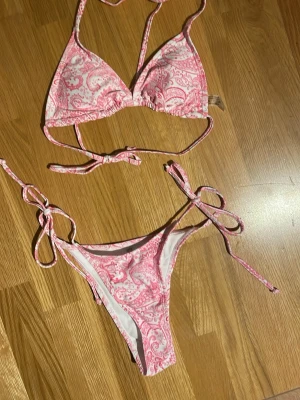 Rosa och vit bikini med knyt - Säljer en snygg bikini i rosa och vitt med paisleymönster. Överdelen har trekantsform och knyt i nacke och rygg, medan underdelen har knyt i sidorna. Perfekt för sommaren och strandhäng! Säljer då jag råkade beställa fel storlek. Skickar helst med PostNord😄