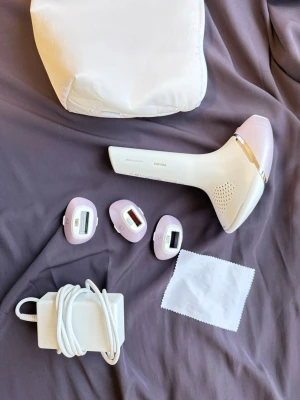 Philips Lumea IPL hårborttagningsapparat - Philips Lumea IPL hårborttagningsapparat i vit och ljusrosa med tre olika behandlingshuvuden för kropp, ansikte och bikinilinje. Medföljer strömadapter, rengöringsduk och praktisk förvaringsväska. Perfekt för smidig och långvarig hårborttagning hemma.