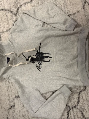 Grå hoodie från Ralph Lauren - Snygg grå hoodie från Ralph Lauren med stor svart logga och tryck framtill. Klassisk känguruficka, huva med vita snören och ribbade muddar. Perfekt för en avslappnad och trendig stil.