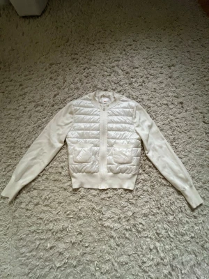 Vit quiltad cardiganjacka - Stilren vit cardiganjacka med quiltad framsida och två fickor. Jackan har ribbade muddar, dragkedja och släta ärmar i stickat material. Perfekt för lager-på-lager och en clean look.