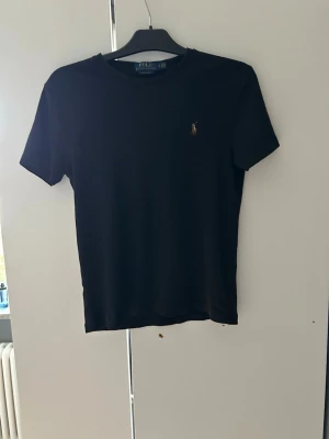 Svart t-shirt från Polo Ralph Lauren - Klassisk svart t-shirt från Polo Ralph Lauren med den ikoniska broderade loggan på bröstet. Storleken är Small-petite vilket innebär att den är lite kortare i armar och längd än en vanlig T-shirt i storlek S