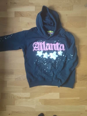 Svart Atlanta hoodie med tryck - Cool svart hoodie från Sprayground med stor rosa 'Atlanta'-text och vita stjärnor på bröstet. Hoodien har huva och är tillverkad i 100% bomull. Unik streetstyle med detaljerade tryck och stänk-effekter på ärmarna.