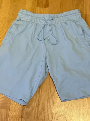 Ljusblå shorts med snörning - Säljer ett par ljusblå shorts med snörning i midjan och sidofickor. Perfekta för sommaren och tillverkade i mjukt bomullsmaterial för maximal komfort. Enkel och stilren design som passar till det mesta.