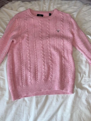 Rosa kabelstickad tröja från Gant - Säljer en rosa kabelstickad tröja från Gant med rund halsringning och broderad logga på bröstet. Tröjan har ribbstickade muddar vid ärmslut och nederkant, och är tillverkad i mjukt material som känns skönt mot huden.