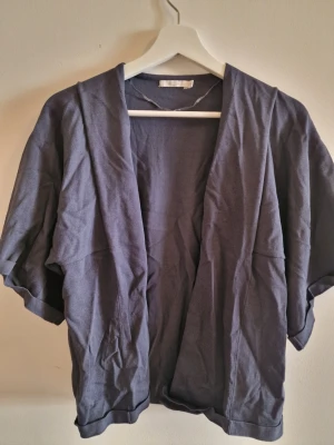 Mörkblå cardiganjacka från Wera - Snygg mörkblå cardiganjacka från Wera med öppen front och avslappnad passform. Koftan har korta ärmar och är tillverkad i ett mjukt tyg som känns skönt mot huden. Perfekt för lager-på-lager och enkel att styla till olika outfits.