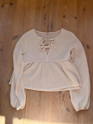 Krämvit blus med knyt i halsen - Söt krämvit blus med långärmad design och knytning vid halsen. Toppen har en lätt textur i tyget och en lös passform nedtill. Perfekt för en trendig och avslappnad look.
