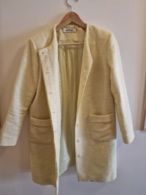 Ljusgul kappa från Rodebjer - Stilren ljusgul kappa från Rodebjer i strukturerat tyg. Rak modell med rundad halsringning, stora fickor framtill och knäppning med dolda knappar. Perfekt för dig som gillar minimalistisk och modern design.