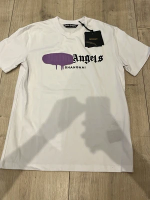 Vit Palm Angels t-shirt Shanghai - Säljer en vit t-shirt från Palm Angels med trycket 'Shanghai' och ett lila grafiskt motiv över bröstet. T-shirten har korta ärmar, rund halsringning och är gjord i mjuk bomull. Perfekt för dig som gillar streetwear och vill sticka ut.