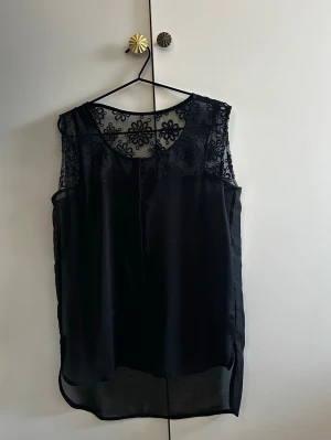 Svart blommig spetslinnetopp - Snygg svart ärmlös linnetopp med transparent mesh och broderade blommor upptill. Toppen har rund halsringning och längre bakstycke för en modern look.             Hör av dig vid frågor!☺️