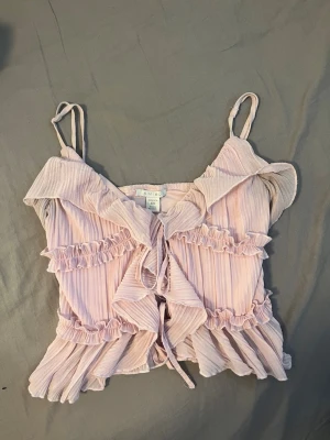 Ljusrosa volangtop från Amisu - Supersöt ljusrosa linnetopp från Amisu med tunna axelband, volangdetaljer och knyt framtill. Toppen har en ribbad struktur och är perfekt för sommarens alla tillfällen. Luftigt material och romantisk vibe.