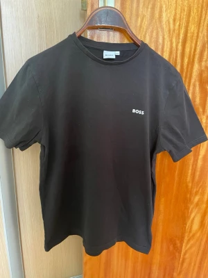 Svart BOSS t-shirt i bomull - Snygg svart t-shirt från BOSS med diskret logga på bröstet. Klassisk rund halsringning och korta ärmar. Tillverkad i mjuk bomull för en skön känsla och enkel stil som funkar till allt.Storlek 14-S. Passar personer som är cirkus 150-155cm. Väldigt fint skick knappast använd. Inga fläckar eller defekter. 