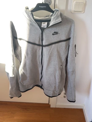 Grå zip hoodie från Nike - Snygg grå hoodie från Nike med dragkedja framtill och svarta detaljer. Klassisk Nike-logga på bröstet och fickor på sidorna. Tillverkad i mjukt material som passar perfekt till vardagslooken. Svart rand går tvärs över bröstet och ärmarna.