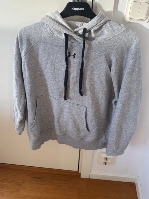 Grå hoodie från Under Armour - Snygg grå hoodie från Under Armour med svart logga på bröstet och svarta dragsnören i huvan. Hoodien har en stor magficka och är tillverkad i mjukt, fleeceliknande material som känns riktigt skönt mot huden. Perfekt för chill eller träning.