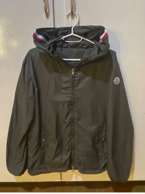 Svart vindjacka från Moncler - Snygg svart vindjacka från Moncler med huva och dragkedja framtill. Jackan har Moncler-logga på bröstet och detaljer i rött, vitt och blått på insidan av huvan. Två fickor med knappstängning och elastiska muddar vid ärmslut. Skicka gärna prisförslag och skriv om ni vill ha mer bilder!