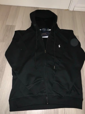Svart hoodie från Polo Ralph Lauren - Svart hoodie från Polo Ralph Lauren med dragkedja, justerbar huva och den klassiska vita logon broderad på bröstet. Tillverkad i mjukt bomullsmaterial och har fickor framtill. Perfekt för en chill och stilren look.