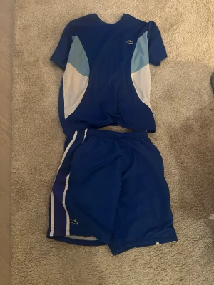 Blått Lacoste träningsset - Säljer ett blått träningsset från Lacoste med t-shirt och shorts. T-shirten har ljusblå och vita paneler på sidorna och en liten Lacoste-logga på bröstet. Shortsen har matchande blå färg med vita och lila detaljer samt Lacoste-logga på benet. 