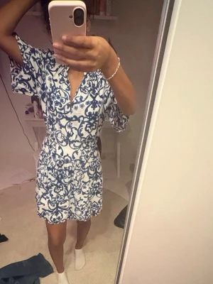 Vit och blå kortklänning med mönster - Kortklänning i vitt med blått, dekorativt mönster. Klänningen har v-ringning, korta vida ärmar och markerad midja. Perfekt för sommarens varma dagar och har en avslappnad men ändå stilren look.