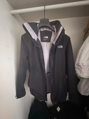 Svart vindjacka från The North Face - Snygg svart vindjacka från The North Face med vit logga på bröstet och ryggen. Jackan har huva, dragkedja framtill och justerbara muddar. Materialet är lätt och vindtätt, perfekt för blåsiga dagar. Enkel och stilren design som passar till det mesta.