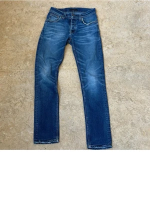 Blå jeans från Nudie Jeans - Säljer ett par klassiska blå jeans från Nudie Jeans med raka ben och snygga slitningar framtill. Jeansen har fem fickor, kontrastsömmar och detaljerade bakfickor med Nudie-loggan. Tillverkade i bomull för en skön och hållbar känsla.