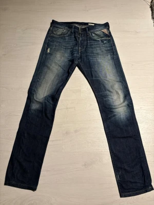 Replay Jeans - Säljer ett par mörkblå jeans med raka ben och snygga slitningar framtill. Jeansen har klassisk femficksdesign och kontrastsömmar. Materialet är denim i bomull och de har en cool, något tvättad look. De har några mindre slitsar efter användning men inget som påverkar helhetsintrycket.                                                            Vid frågor är det bara att höra av sig!