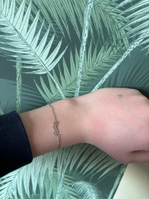 Saint Laurent armabnd - Snyggt och stilrent armband i silverfärg från Yves Saint Laurent med den ikoniska YSL-logotypen som hänge. Tunt kedjearmband med diskret design och liten bricka med ingraverad logga. Perfekt accessoar för dig som gillar exklusiva detaljer.