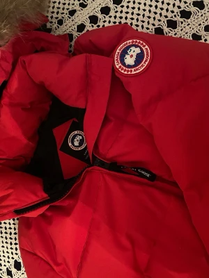 Röd dunjacka från Canada Goose - Säljer en röd dunjacka från Canada Goose med huva och avtagbar pälskant. Jackan har dragkedja framtill, två sidofickor och klassisk Canada Goose-logga på ärmen. Köpt av en annan och därav inget kvitto eller dustbag. Storlek L/G men passar från S-L