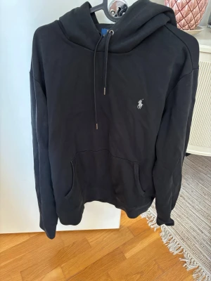 Svart hoodie från Polo Ralph Lauren - Klassisk svart hoodie från Polo Ralph Lauren med broderad logga på bröstet. Hoodien har huva med dragsko och en stor magficka framtill. Tillverkad i mjukt bomullsmaterial och har en avslappnad passform som passar perfekt till streetwear-stilen.