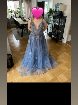 Blå långklänning med spetsdetaljer -  Långklänning med tunna axelband, djup v-ringning och vackra spetsapplikationer i silver. Klänningen har snörning i ryggen och ett lager av tunn tyll.Klänningen är skräddarsydd men passar förmodligen alla strl från xs-l, eftersom att snörningen är justerbar. Det är en större kupa på modellen och jag har 85 c och den passar perfekt. Klänningen med klackar för mig var lite lång och jag är 157 cm lång, därför gissar jag att den passar någon runt 165 cm-170. Den är helt ny och har endast provats.