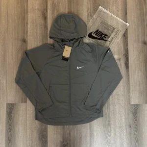 🩶Grå vindjacka med huva från Nike🩶 - Snygg grå vindjacka från Nike med huva och dragkedja framtill. Jackan har två sidofickor och en diskret vit Nike-logga på bröstet. Tillverkad i lätt och vindtätt material, perfekt för blåsiga dagar. Enkel och stilren design som passar till det mesta. Tar emot rimliga bud. Helt ny oanvänd.