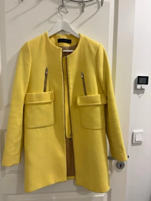Gul kappa från Zara Woman - Stilren gul kappa från Zara Woman med rund halsringning, två stora fickor framtill och dragkedjor som detaljer. Kappan har en rak passform och är fodrad i beige. Perfekt för dig som vill sticka ut med färg i din outfit.