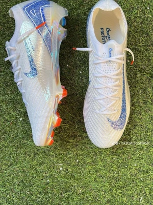 Vita Nike Mercurial fotbollsskor - Snygga vita Nike Mercurial fotbollsskor med skimrande blått swoosh och holografiska detaljer. Skorna har en stickad ovandel och fasta dobbar i orange och silver för bästa grepp på planen. Perfekta för dig som vill sticka ut på fotbollsplanen.