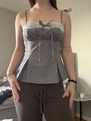 Grå linnetopp med spets och rosett - Supersöt grå linnetopp med tunna axelband som knyts på axlarna. Toppen har vit spetskant upptill och en dekorativ rosett framtill, även utsvängd❤️