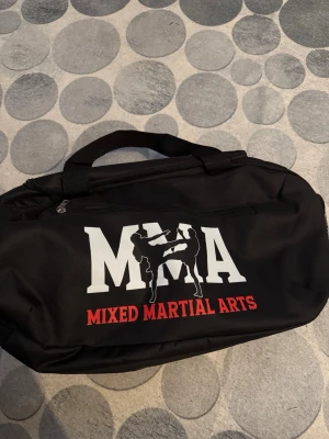 Svart MMA träningsväska - Svart träningsväska med tryck i vitt och rött där det står 'MMA Mixed Martial Arts' och en siluett av två fighters. Väskan har dragkedja och bärhandtag, perfekt för gym eller kampsportsträning. Tillverkad i slitstarkt tygmaterial.