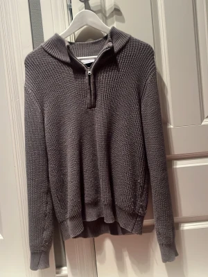 Grå stickad tröja med halv dragkedja - Mysig grå stickad tröja med halv dragkedja framtill och krage. Tröjan har lång ärm, ribbade muddar och nederkant samt en avslappnad passform. Perfekt för lager på lager och enkel att matcha med olika stilar.
