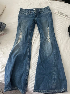 Gina 14+ jeans - Säljer ett par blå bootcut jeans med slitningar på framsidan och klassiska fem fickor. Jeansen har låg midja och vida ben, perfekt för en avslappnad och trendig look. Materialet är denim i bomull och de har snygga detaljer på bakfickorna.