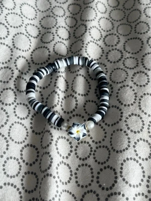 Svartvitt pärlarmband med blomma - Unikt armband med svarta och vita platta pärlor, två vita runda pärlor och en snygg blomformad pärla i mitten med svart, vitt och lite gult. Perfekt för dig som gillar coola och kreativa accessoarer.