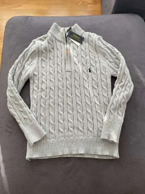 Stickad tröja från polo - Ljusgrå kabelstickad tröja från Polo Ralph Lauren med halv dragkedja och hög krage. Tröjan har ribbade muddar och nederkant samt den klassiska broderade loggan på bröstet. Tillverkad i mjukt material som passar perfekt till höst och vinter.