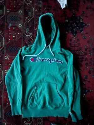 Grön Champion hoodie  - Snygg grön hoodie från Champion med stor logga över bröstet. Klassisk känguruficka framtill och vita dragsnören i huvan. Perfekt för dig som gillar streetwear och vill ha en bekväm och stilren look.