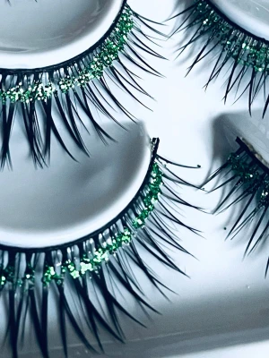 Prisförslag- 1 par Långa lösögonfransar med grön glitter - Snygga lösögonfransar med intensiv volym och längd. Fransarna är svarta med en rad av grönt glitter längs fransroten som ger en unik och iögonfallande look. Perfekta för dig som vill sticka ut och addera extra effekt till din ögonmakeup.